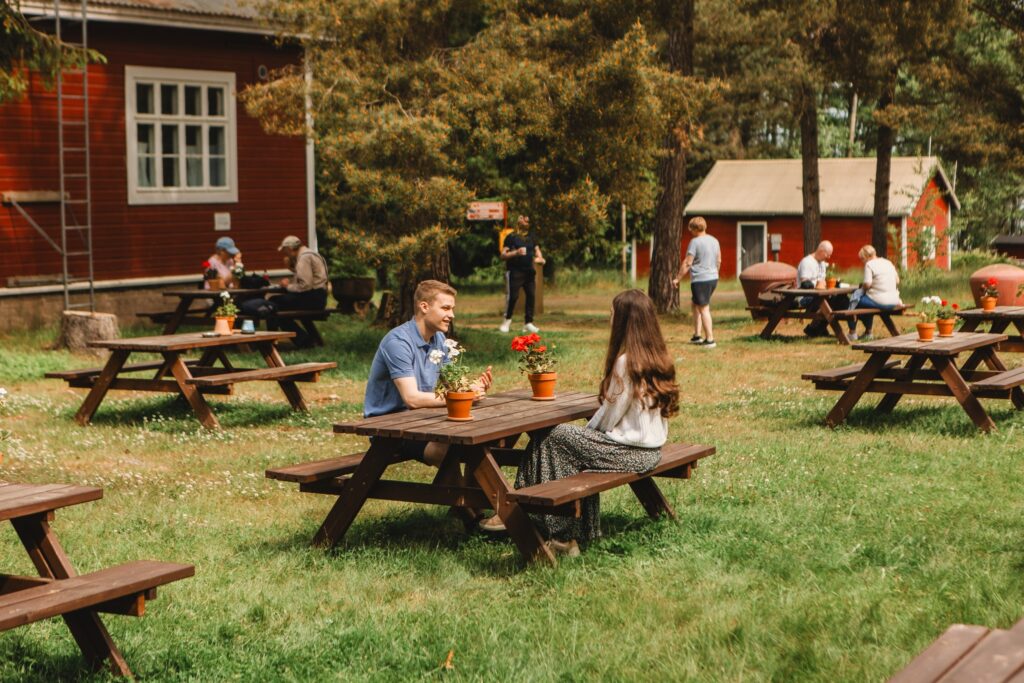 Ihmisiä viettämässä aikaa saaristokohteen piha-alueella piknikpöytien äärellä.
