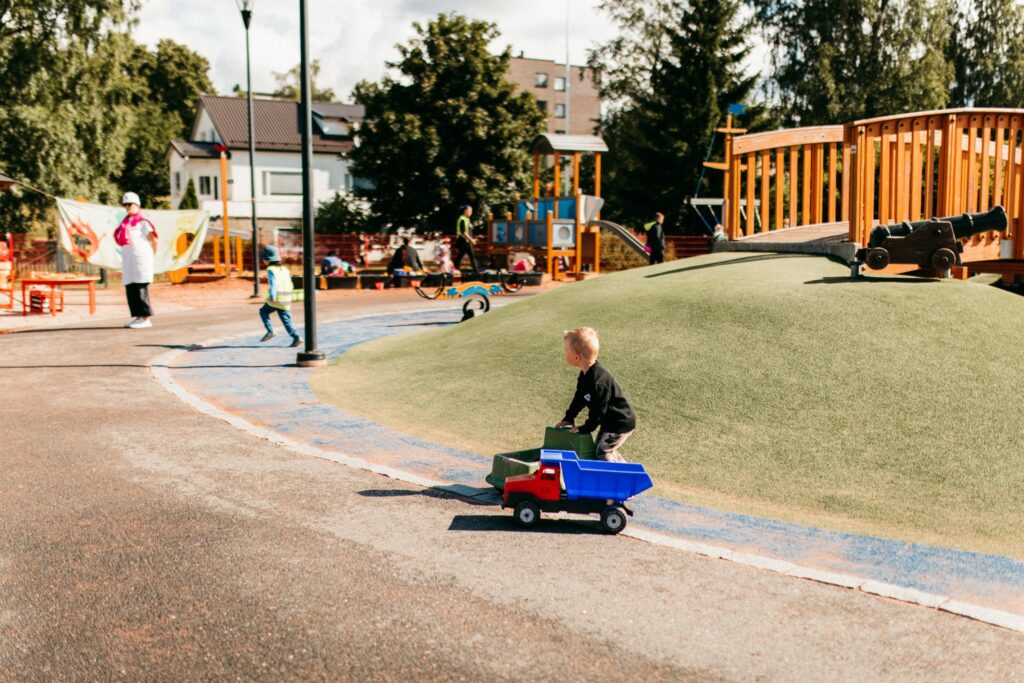 Lapsi leikkii leikkipuistossa vetäen leluautoa muiden lasten ja aikuisten ollessa taustalla.
