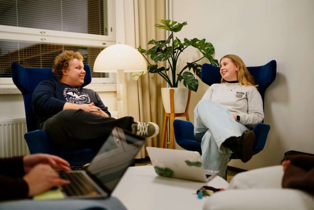 Neljä nuorta opiskelijaa keskustelee rennosti nojatuoleissa sisätilassa kannettavan tietokoneen äärellä.