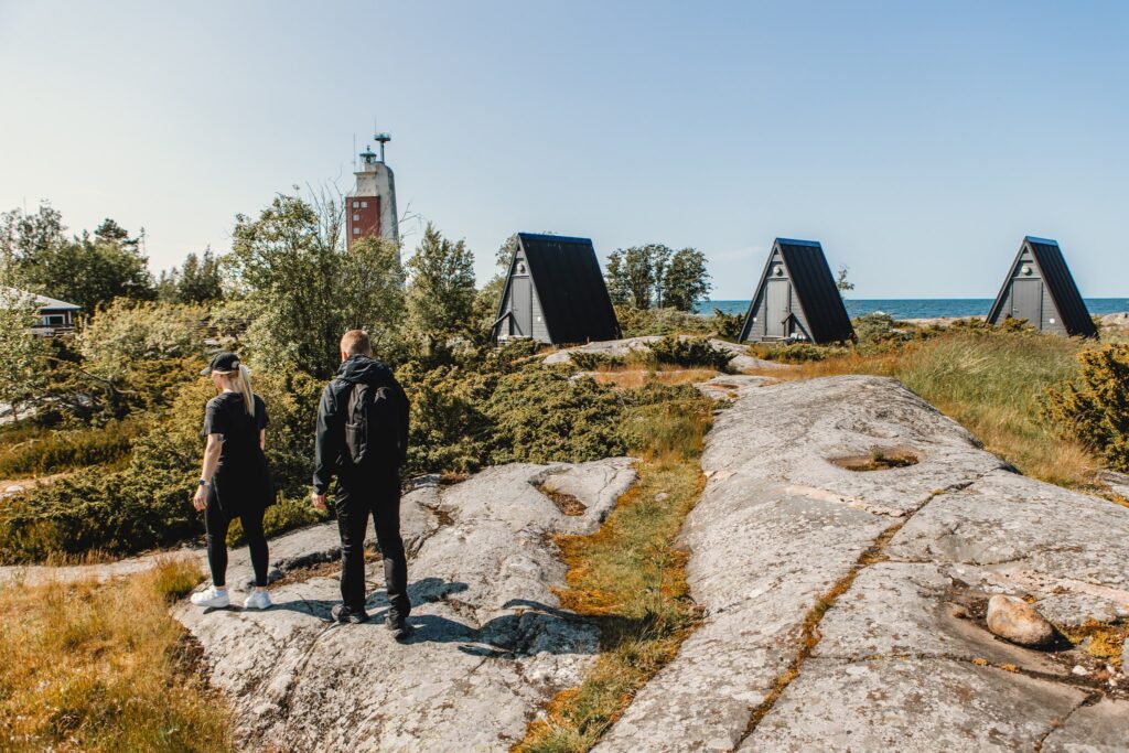 Kaksi retkeilijää tutustuu saaristokohteeseen, jossa kalliomaisema, majoitusmökit ja majakka muodostavat merellisen matkailuympäristön.